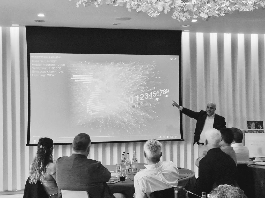 ehab shanti in a suit stands beside a large projection screen, pointing to a dynamic data-visualization slide filled with network-like dots and the numbers 0–9. Several attendees sit around round tables in the foreground, watching attentively in a modern conference room with vertical wall panels.
